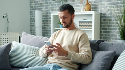 Joyful bearded man is using smartphone texting having fun smiling in house alone relaxing on sofa enjoying leisure time with new technologies. Youth and gadgets concept.