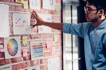 Asian man creative director designer looking at data chart and find idea on brickwall at modern office.brainstroming creative ideas concpet.