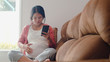 © tirachard - Young Asian Pregnant woman using mobile phone search pregnancy information. Mom feeling happy smiling positive and peaceful while take care her child lying on sofa in living room at home concept.