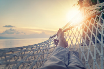 Wall Mural - Female legs in a hammock on a background of the sea, palm trees and sunset. Vacation concept, point of view
