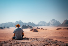 Guy Sitting On A Rocky Outcrop In The Desert Of Wadi Rum With Mountains In The Background