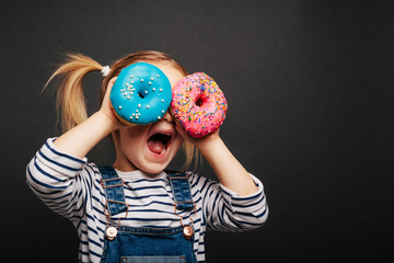 Happy cute girl is having fun played with donuts on black background wall.