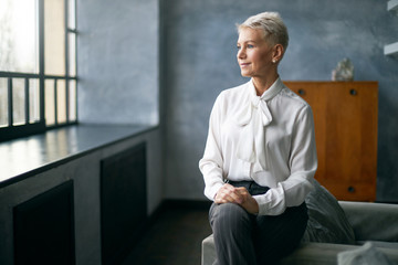 Indoor image of confident beautiful middle aged female psycologist wearing white blouse sitting on couch in her office, having calm facial expression, looking through window, waiting for client