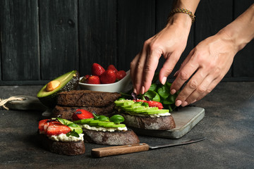 Wall Mural - Toast or sandwich with avocado, cheese, strawberries, herbs and seeds on a dark background. Female hands serve a dish. An idea for bruschetta or for a healthy snack. Healthy vegan breakfast.