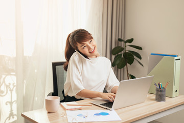Fototapete - Confident Asian woman talking on her mobile phone and using laptop while sitting at desk and working from home. Home office.