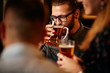 © chika_milan - Handsome bearded caucasian young man with eyeglasses standing in pub with friends and drinking fresh beer.