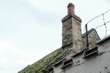 Very Old, Domestic Home Showing A Moss Covered Roof Together With A Single, Brick Built Chimney. Also In View Is A Barbed Wire Fence To Stop People Climbing Over The Low Roof Line.