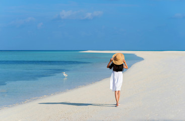 Back of young Asian girl walking on white sand beach with clear blue sea and sky. Teenager girl wearing sun straw hat, white dress enjoy vacation. Outdoor summer vacation travel concept, copy space.