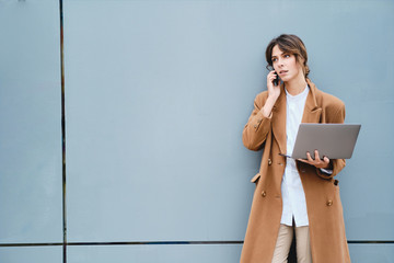 Young attractive businesswoman in coat with laptop thoughtfully talking on cellphone outdoor