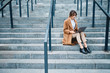 © Anton - Young attractive stylish businesswoman in coat confidently working on laptop on stairs outdoor
