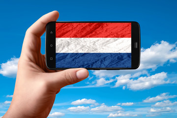 Holland flag on the phone screen. Smartphone in hand shows a flag on a background of the sky with clouds.