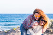 © simona - Couple of adult nice caucasian women play together carrying eachother with lot of fun and laughs - people laughing in outdoor leisure activity - ocean in background for vacation lifestyle