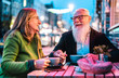 Hipster retired senior couple in love enjoying cappuccino at outdoor cafeteria - Joyful elderly lifestyle concept with wife and husband having fun at bar cafe restaurant - Vivid neon lights filter