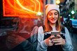 © Westend61 - Smiling young woman standing next to neon light using her smartphone at night