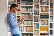© Westend61 - Smiling young man standing in front of bookshelves at home using digital tablet