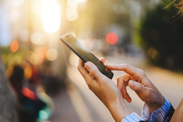 Cropped image of female person using modern mobile phone device with touch screen outdoors on city bokeh lights background,