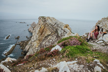 Adult Woman In Casual Clothes Looking Away And Enjoying Views While Sitting And Hugging Border Collie Dog On Rocky Shore Against Gray Waves Under Cloudy Sky In Asturias In Summer