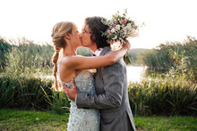 Stylish Newly Married Couple In Wedded Suits Embracing And Kissing With Green Plants And Mere On Sunny Day