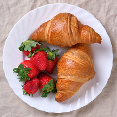 View on two fresh croissants with ripe strawberries on white plate and natural linen napkin background. Classic French European breakfast. Warm croissants and red berries.