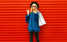 Attractive Young Woman With Shopping Bags, Stylish Female Model Showing Peace Gesture Wearing Blue Faux Fur Coat, Round Hat And Sunglasses Over Red Wall Background