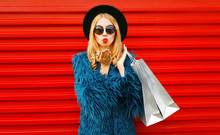 Attractive Young Woman With Shopping Bags Blowing Red Lips Sending Sweet Air Kiss, Stylish Female Model Wearing Blue Faux Fur Coat, Round Hat And Sunglasses Over Red Wall Background