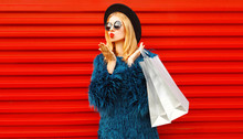 Side View Beautiful Woman With Shopping Bags Blowing Red Lips Sending Sweet Air Kiss, Stylish Female Model Wearing Blue Faux Fur Coat, Round Hat And Sunglasses Over Red Wall Background