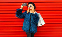 Attractive Young Smiling Woman Taking Selfie Picture By Smartphone With Shopping Bags, Stylish Female Model Wearing Blue Faux Fur Coat, Black Round Hat And Sunglasses Over Red Wall Background