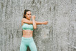 © annanahabed - Outdoor portrait of young beautiful fit woman stretching arms, wearing green activewear, athlete model posing next to grey urban wall background, sport fashion
