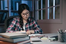 Young Asian Chinese Woman In Eyeglasses Studying Late At Night At Home. Hard Working Girl Student Reading Textbook And Review Information For Exam In Bedroom. Education And Deadlines Concept