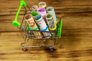 Shopping cart with money from different countries: euros, us dollars, ukrainian hryvnia, russian rubles, egyptian pounds on wooden background