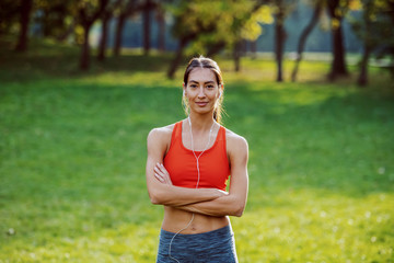 Wall Mural - Fit healthy slim smiling caucasian brunette in sportswear and with ponytail standing on meadow with arms crossed. In ears are earphones. Sunny day in nature.