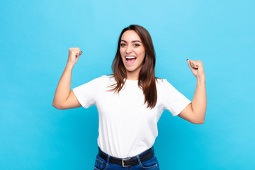 young pretty woman feeling happy, satisfied and powerful, flexing fit and muscular biceps, looking strong after the gym against blue wall