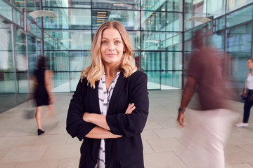 Portrait Of Businesswoman With Crossed Arms Standing In Lobby Of Busy Modern Office