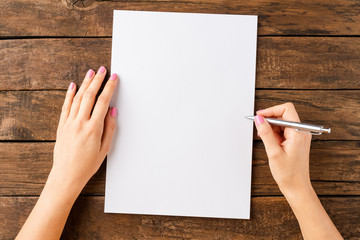 Woman’s hands writing with pen over blank white paper sheet on wooden table. Top view