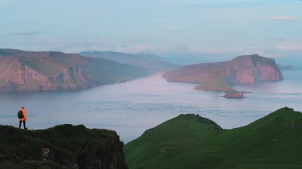 Wall Mural - Lonely tourist in red jacket looking over majestic Alaberg cliffs on Mykines island, Faroe Islands. UHD 4k video