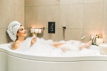 Happy Beautiful Woman Relaxing In A Bubble Bath Tub, Holding Glass Of Wine, White Towel On The Head