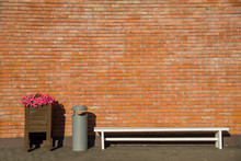 White Bench, Recycle Bin And Pink Flowers In A Wooden Box On A Background Of An Old Red Orange Brick Wall