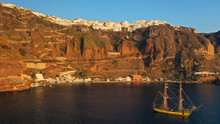 Aerial Drone Photo Of Wooden Sail Boat Docked Near Old Port Of Santorini Island In Deep Blue Sea Just Below Village Of Fira, Cyclades, Greece