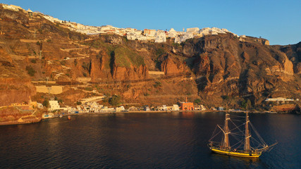 Aerial drone photo of wooden sail boat docked near old port of Santorini island in deep blue sea just below village of Fira, Cyclades, Greece