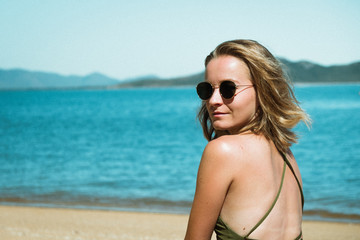 Wall Mural - Young attractive girl with sunglasses sitting on the beach, getting tanned, looking to the camera