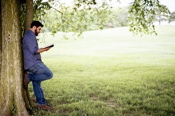 Male leaned against a tree while reading the bible with a blurred background