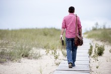 Male Walking On A Wooden Pathway Carrying His Bag And Holding The Bible With A Blurred Background