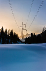 Power pole and power lines at sunset in a valley in winter. Fresh snow in foreground, forest and mountains in background with yellow hue