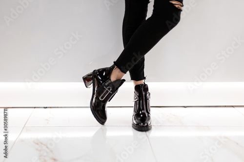 Fotomural Closeup of female legs in black jeans in lacquered black boots on silver heels near a white wall in the room