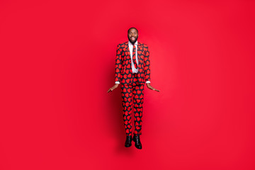 Full size photo of crazy dark skin hipster guy jumping high demonstrating cool modern trendy look wear stylish hearts pattern suit blazer shirt necktie tie trousers isolated red color background