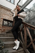 Urban fashion model of a cute young woman in spring-autumn clothes in trendy boots posing in the city on a staircase with old wooden railing. Attractive girl in a stylish outerwear stands outdoors.