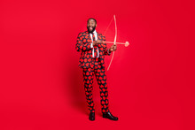 Full Body Photo Of Funny Dark Skin Man Shooting Bow Love Arrow Amour Cupid Good Couple Desire Wedding Wear Hearts Pattern Suit Shirt Necktie Tie Shoes Isolated Red Color Background