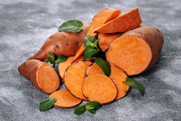 Wall Mural - Sweet Potato Slices Decorated Mint Green Leaves. Heap of Nourishing Orange Batata with Flavored Plant Ingredients for Healthy Lunch. Natural Nutrition Isolated on Grey Kitchen Table