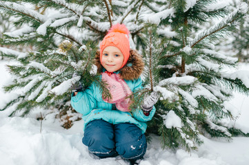 Wall Mural - Happy little girl in winter clothes sitting outdoor under spruce tree covered with snow