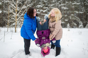  Two adult positive woman holding little girl legs upside down in winter snowy field.
